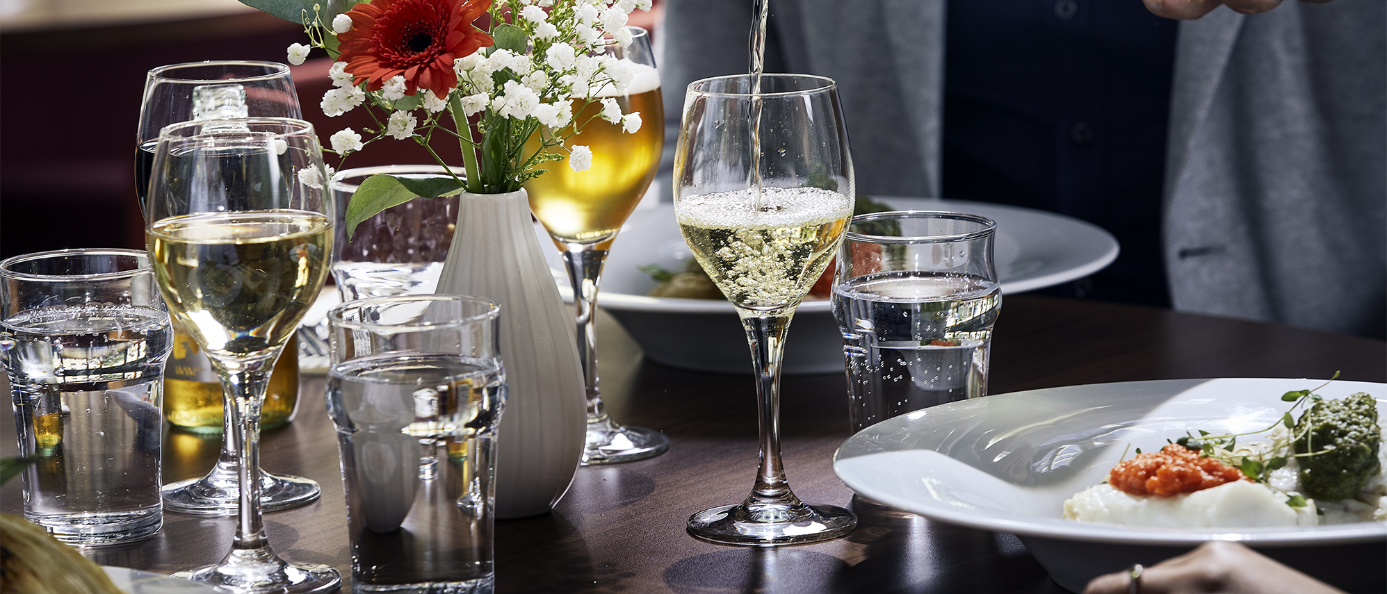 Dukat bord med glas, tallrikar och blommor, servitris häller vin i ena glaset.